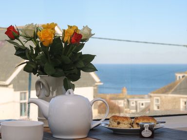 A table with a white teapot bowl plate with scones and a vase of roses by a window with a sea view at The Beach Hut in St Ives