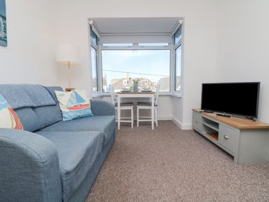 A living room with a blue sofa a floor lamp a dining table with two chairs and a television on a cabinet at The Beach Hut in St Ives
