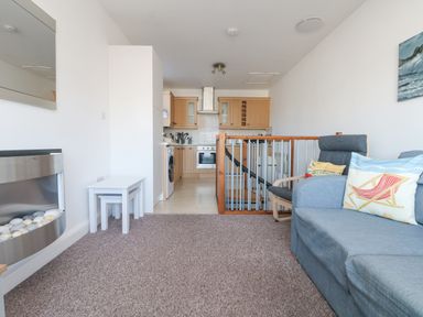 A living room with a blue sofa and chair next to a railing with a kitchen in the background at The Beach Hut in St Ives