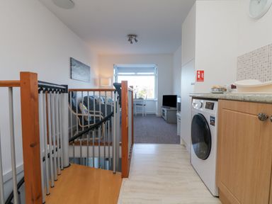 A kitchen area with a washing machine and wooden cabinets next to a staircase and a living room with a couch and television at The Beach Hut in St Ives