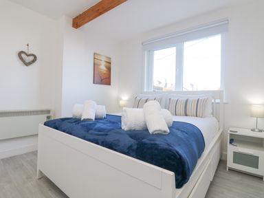 A bedroom with a white bed, blue blanket, rolled towels, striped pillows, nightstands, lamps, and window at The Beach Hut in St Ives