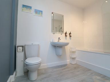 A bathroom with a toilet, wall-mounted sink, mirror, bathtub with a shower, and wall art at The Beach Hut in St Ives
