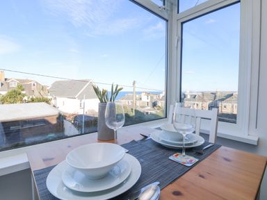 A dining table set with white bowls plates and wine glasses by a window overlooking rooftops and the sea at The Beach Hut in St Ives