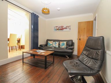 A living room with a black leather sofa and chair a wooden coffee table with coasters and a doorway with yellow chairs visible at Weybury Cottage Brewers Quay Harbour