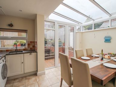 A dining area with a wooden table set with plates and glasses next to a kitchen with a washing machine and a door leading outside at Weybury Cottage Brewers Quay Harbour