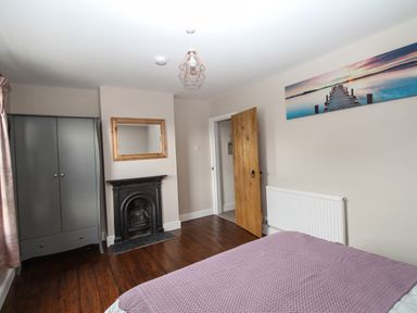 A bedroom with a bed covered with a purple blanket a wooden floor a black fireplace a grey wardrobe a wooden door and a landscape photo on the wall at Weybury Cottage Brewers Quay Harbour
