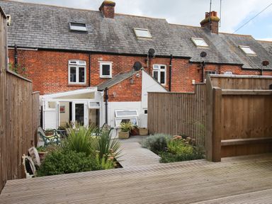 A backyard with a wooden deck a garden path and plants next to a row of red brick houses with skylights and a glass door at Weybury Cottage in Brewers Quay Harbour