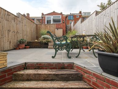 A backyard patio area with stone steps leading to a paved seating area with green metal chairs and table surrounded by wooden fences and plants at Weybury Cottage Brewers Quay Harbour