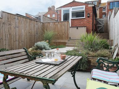An outdoor patio with wooden table and benches with cushions surrounded by plants and wooden fences at Weybury Cottage Brewers Quay Harbour