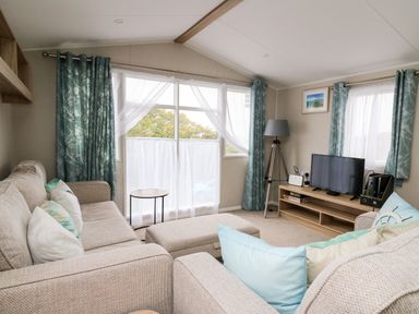 A living room with beige sofas cushions a small round table a TV on a wooden stand and blue patterned curtains at Ystwyth 26 in Borth