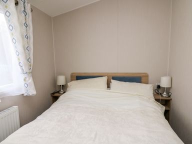 A bedroom with a double bed two pillows two bedside tables with lamps and a window with patterned curtains at Ystwyth 26 in Borth