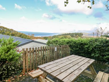 A wooden picnic table with benches on a patio surrounded by bushes and a wooden fence with a view of the sea and hills at Ystwyth 26 in Borth