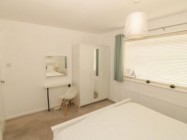A bedroom with a white bed a white wardrobe a chair a small desk a mirror and a window with blinds and green curtains at Seaside retreat in Porthmadog