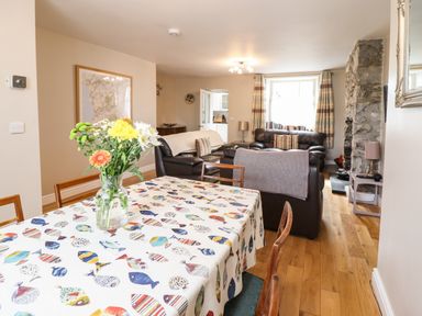 A dining area with a table covered in a fish-patterned tablecloth and flowers in a vase next to a living room with leather sofas and a stone fireplace at Artro View in Llanbedr