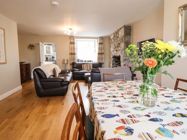 A living and dining area with a wooden table with a floral centerpiece a patterned tablecloth leather sofas a stone fireplace and hardwood floors at Artro View in Llanbedr