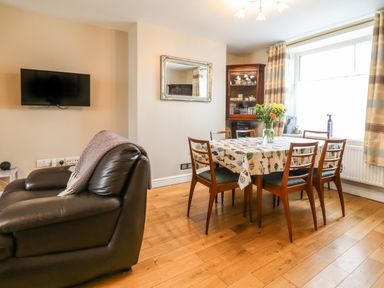 A dining room with a wooden table covered with a fish patterned tablecloth surrounded by six chairs next to a large window and a leather sofa with a blanket on the armrest at Artro View in Llanbedr