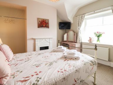 A bedroom with a bed with floral bedding white folded towels a vanity table a wall-mounted tv a painting above a fireplace and a window with curtains and flowers at Artro View in Llanbedr