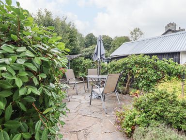 An outdoor stone patio with a round table and four chairs surrounded by greenery and a building with a corrugated metal roof at Artro View in Llanbedr