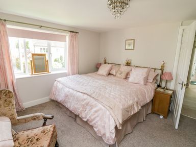 A bedroom with a large bed pink curtains wooden bedside table and floral armchair at Crooked Well in Timsbury Nr Bath