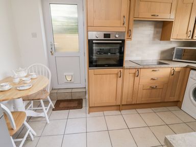 A kitchen with wooden cabinets a built in oven a microwave a washing machine a round wooden table with tea set and two white chairs at Crooked Well in Timsbury Nr Bath