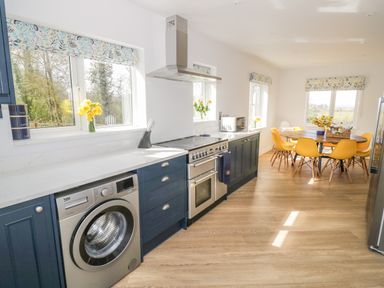 A kitchen with blue cabinets, stainless steel oven and washing machine, wooden dining table with yellow chairs, and windows with floral blinds at Orchard House Risbury near Leominster