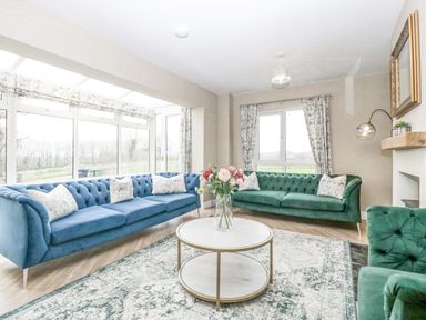 A living room with blue and green sofas a round coffee table with flowers and large windows at Orchard House in Risbury near Leominster