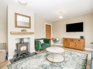 A living room with a green armchair a round coffee table with flowers a wooden cabinet a wall-mounted TV and a fireplace at Orchard House in Risbury near Leominster
