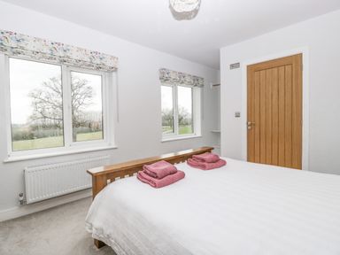 A bedroom with a wooden bed frame white bedding folded towels on the bed two windows with patterned blinds a radiator under one window and a wooden door at Orchard House in Risbury near Leominster