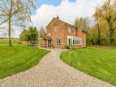 A brick house with a gravel pathway and green lawn surrounded by trees at Orchard House in Risbury near Leominster