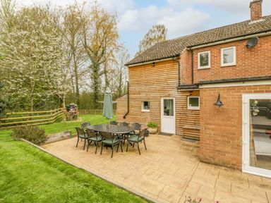 A patio with a table and chairs next to a brick and wood exterior of a house with a garden and trees at Orchard House in Risbury near Leominster