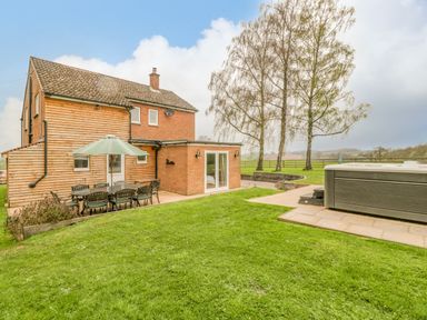 A backyard with a dining table and umbrella beside a house and a hot tub near trees at Orchard House in Risbury near Leominster