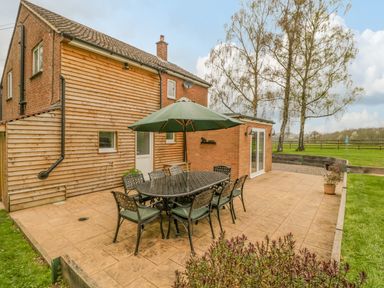 An outdoor patio with a dining table and chairs under an umbrella next to a brick house with wooden siding and trees in the background at Orchard House in Risbury near Leominster