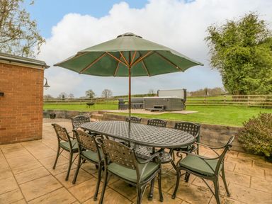 An outdoor patio with a metal table and chairs under a large green umbrella next to a lawn with hot tub and fencing at Orchard House in Risbury near Leominster