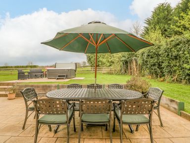 An outdoor patio with a metal table and chairs under a large green umbrella and a hot tub on a raised platform at Orchard House in Risbury near Leominster