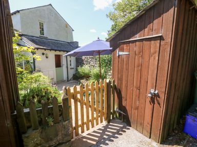 A garden area with a wooden gate a wooden shed a patio umbrella and a house with a green door at St Peters Cottage in Milnthorpe