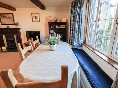A dining room with a table covered with a cloth and wooden chairs a fireplace a television a bookshelf and a window bench at St Peters Cottage in Milnthorpe