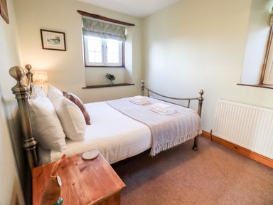 A bedroom with a metal bed, wooden bedside table, two windows, and folded towels at St Peters Cottage in Milnthorpe