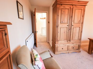 A room with a wooden wardrobe a beige chair with floral cushion and an open door showing a hallway at St Peters Cottage in Milnthorpe