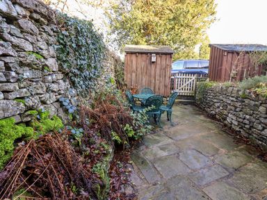 A stone patio with green metal table and chairs between stone walls and wooden sheds at St Peters Cottage in Milnthorpe