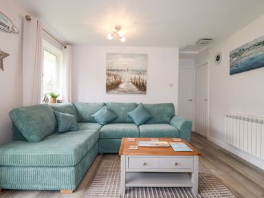 A living room with a teal sectional sofa a wooden coffee table and coastal paintings at Blue Sky Cottage Rosecraddoc Lodge near Liskeard