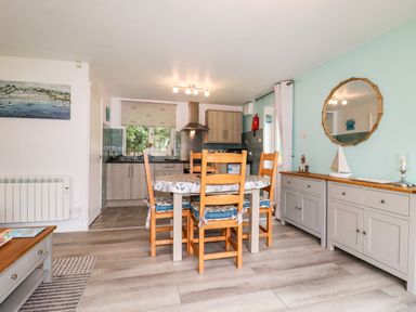 A dining area with a wooden table and chairs next to a kitchen and a sideboard with a round mirror at Blue Sky Cottage in Rosecraddoc Lodge near Liskeard