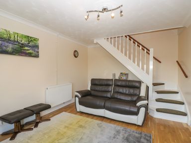 A living room with a leather sofa two stools a wall clock a radiator a staircase and a landscape picture at The Annex in Drybrook