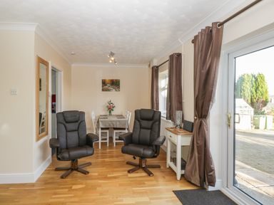 A dining area with a table and chairs two black armchairs wooden flooring and a glass door with curtains at The Annex in Drybrook