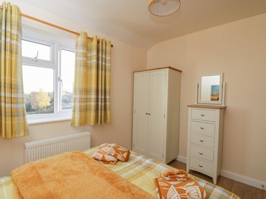 A bedroom with a bed covered in orange and yellow bedding next to a window with yellow plaid curtains and white wardrobe and chest of drawers in the corner at The Annex in Drybrook