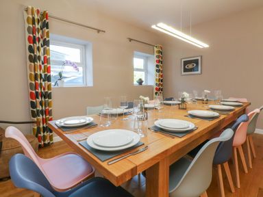 A dining room with a wooden table set with plates glasses and cutlery and multicolored chairs by two windows with patterned curtains at The Old Coach House in Braunton