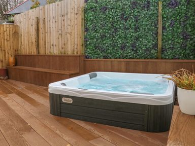 An outdoor hot tub on a wooden deck with a wooden bench and a green leafy wall at The Old Coach House in Braunton