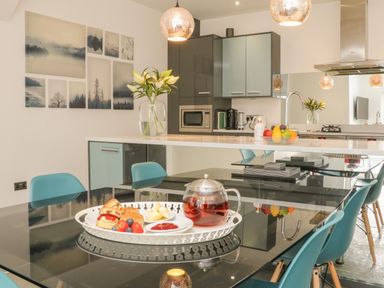 A kitchen and dining area with blue chairs flowers on a countertop and a tray of food and tea on a glass dining table at The Old Coach House in Braunton