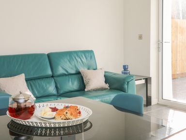 A teal sofa with cushions and a tray with scones and tea on a glass table at The Old Coach House in Braunton