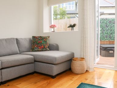A grey sectional sofa with a patterned cushion a wicker basket a window with flowers and peacock feathers and an open door showing a wooden deck with a hot tub at The Old Coach House in Braunton