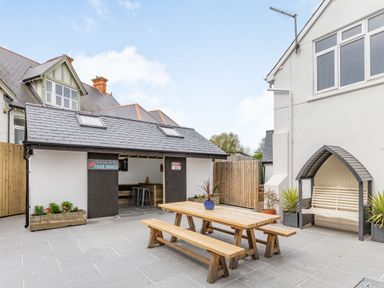 An outdoor patio with a wooden picnic table and bench a covered bench seat and a small building with signs on the doors at The Old Coach House in Braunton